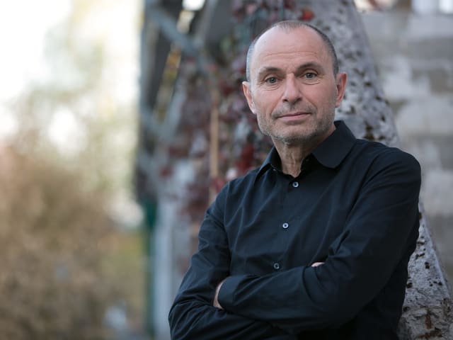Mann (Norbert Gstrein) mit schwarzem Hemd und fast Glatze steht mit verschränkten Händen vor einem Baum. Mann (Norbert Gstrein) mit schwarzem Hemd und fast Glatze steht mit verschränkten Händen vor einem Baum.