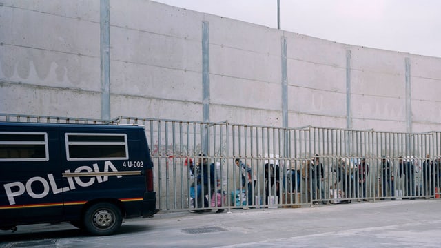 Eine Mauer, vor der ein Polizeiauto steht und Menschen mit Tüten Eine Mauer, vor der ein Polizeiauto steht und Menschen mit Tüten