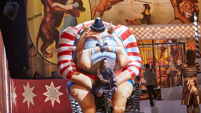 Eine Frau fährt auf der Theaterbühne ein Fahrrad, auf dem ein aufgeblasenes Monster montiert ist. Eine Frau fährt auf der Theaterbühne ein Fahrrad, auf dem ein aufgeblasenes Monster montiert ist.