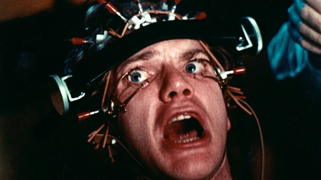 Ein Mann mit gequältem Blick mit einer Apparatur auf dem Kopf, die ihm die Augen offen hält. Ein Mann mit gequältem Blick mit einer Apparatur auf dem Kopf, die ihm die Augen offen hält.