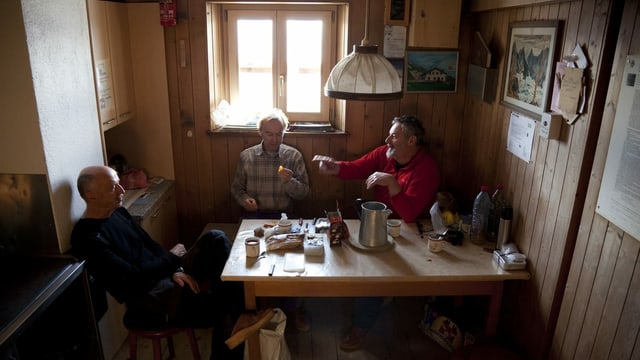 Drei Männer sitzen in einer kleinen Stube an einem Holztisch Drei Männer sitzen in einer kleinen Stube an einem Holztisch
