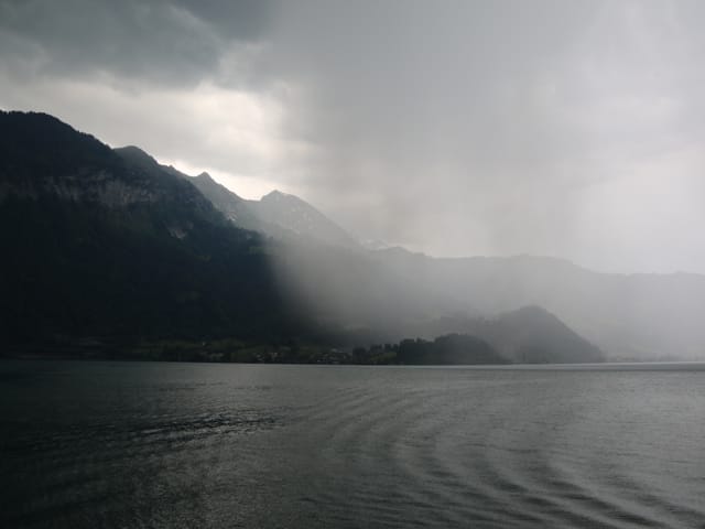 See bei garstigem Wetter. Am anderen Ufer Konturen von schroffen Bergen. Von rechts kommt eine Zone mit Regen, eher weisslich.