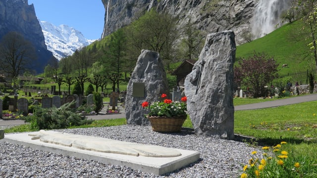 Zwei grosse Steine, eine Betonplatte mit eingravierter Feder, im Hintergrund Berge mit Schnee und ein Wasserfall Zwei grosse Steine, eine Betonplatte mit eingravierter Feder, im Hintergrund Berge mit Schnee und ein Wasserfall