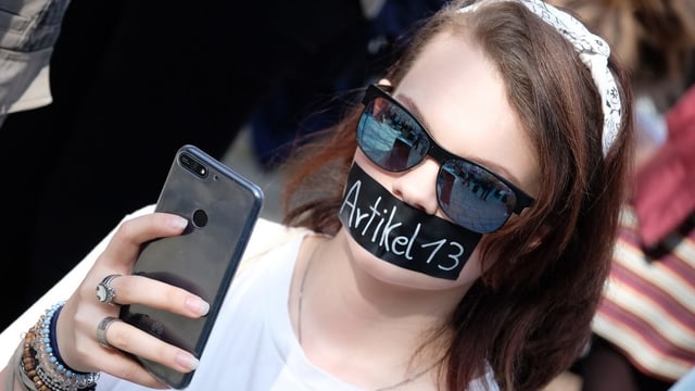 Junge Frau mit Kleber auf dem Mund. Darauf steht Artikel 13. Sie macht ein Selfie. Junge Frau mit Kleber auf dem Mund. Darauf steht Artikel 13. Sie macht ein Selfie.