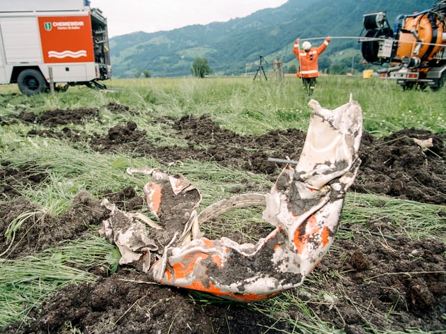 Wrackteil im Gras, hinten ein Feuerwehr- und ein Kanalreinigungsfahrzeug.