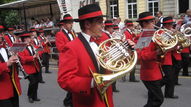 Musicants en giaccas cotschnas e cun chapella, che sunan durant marschar atras ina via da la citad da Cuira. Musicants en giaccas cotschnas e cun chapella, che sunan durant marschar atras ina via da la citad da Cuira.
