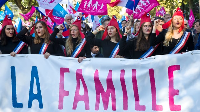 Junge Frauen mit violetten phrygischen Mützen tragen ein Plakat «Familie».