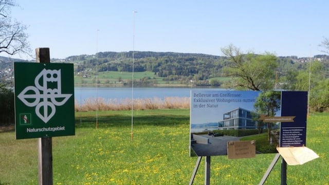 Vor einem See stehen zwei Tafeln am Ufer. Eines zeigt das Symbol des Naturschutz, daneben wirbt ein Plakat für Luxuswohnungen. Vor einem See stehen zwei Tafeln am Ufer. Eines zeigt das Symbol des Naturschutz, daneben wirbt ein Plakat für Luxuswohnungen.