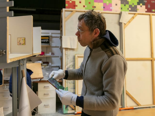 Mann mit kurzen Haaren, Brille und grauem Pullover steht mit einem kleinen Bild in der Hand vor einem Regal. Mann mit kurzen Haaren, Brille und grauem Pullover steht mit einem kleinen Bild in der Hand vor einem Regal.