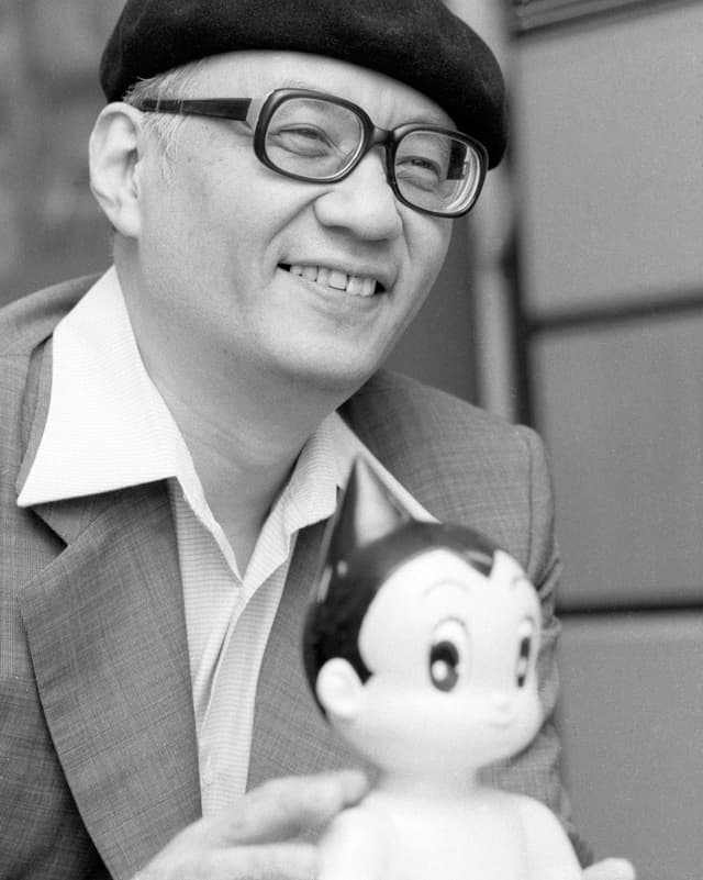 Der lächelnde Osamu Tezuka mit Beret und Brille in einer schwarzweiss Aufnahme, vor ihm steht eine «Astroboy»-Figur. Der lächelnde Osamu Tezuka mit Beret und Brille in einer schwarzweiss Aufnahme, vor ihm steht eine «Astroboy»-Figur.