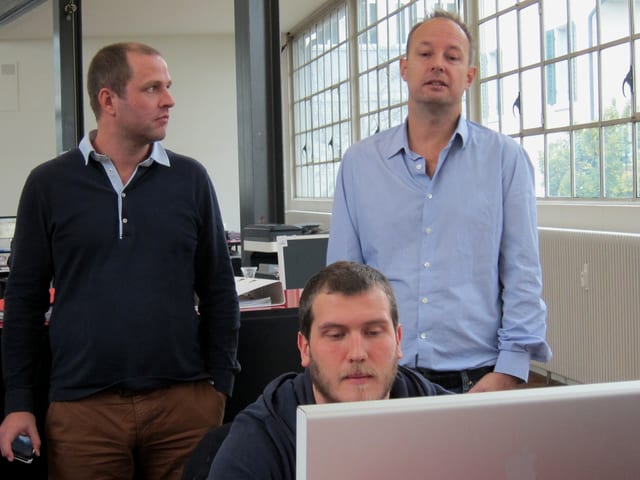 Ein Mann sitzt am Pult vor einem Computer, hinter ihm stehen zwei weitere Männer, von denen einer ebenfalls auf den Bildschirm schaut. Ein Mann sitzt am Pult vor einem Computer, hinter ihm stehen zwei weitere Männer, von denen einer ebenfalls auf den Bildschirm schaut.
