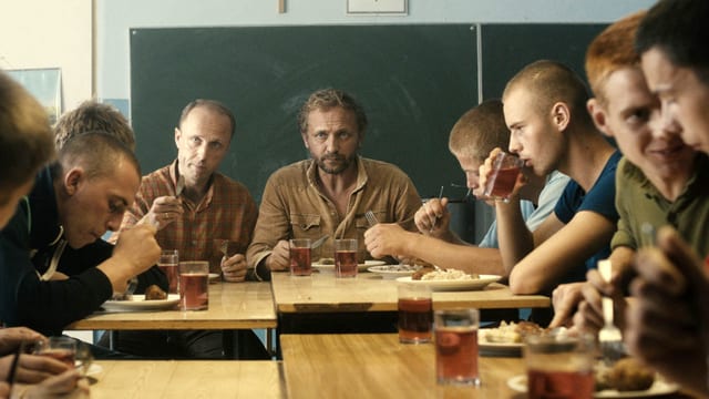 Rund um einen Tisch sind jüngere und zwei ältere Männer zum Essen versammelt. Rund um einen Tisch sind jüngere und zwei ältere Männer zum Essen versammelt.