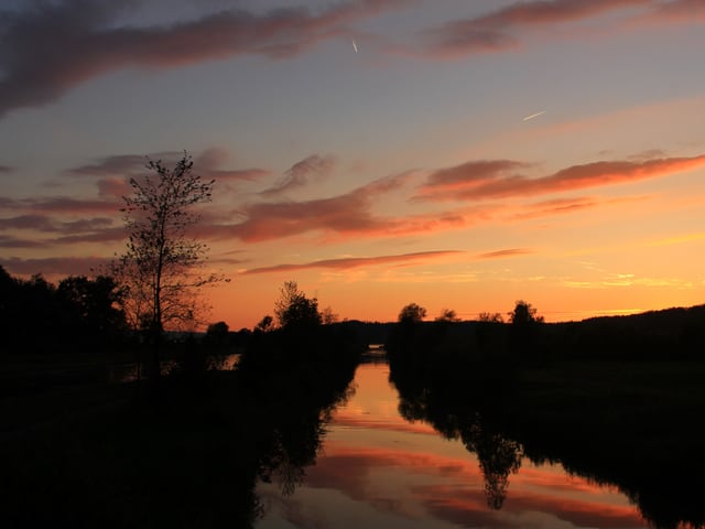 Das Bild ist in rosa und lila Farben gehalten. Man erkennt im Abendlicht nur Konturen von einem Fluss, Bäumen und Feldern. Am Himmel hat es ein paar wenige Wolken.