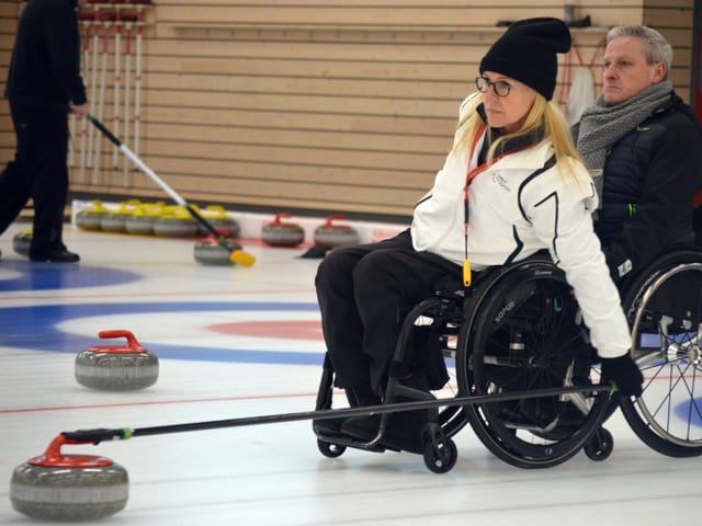 Eine Frau mit Rollstuhl hält eine lange Stange mit einem Curlingstein am Ende in den Händen. Eine Frau mit Rollstuhl hält eine lange Stange mit einem Curlingstein am Ende in den Händen.