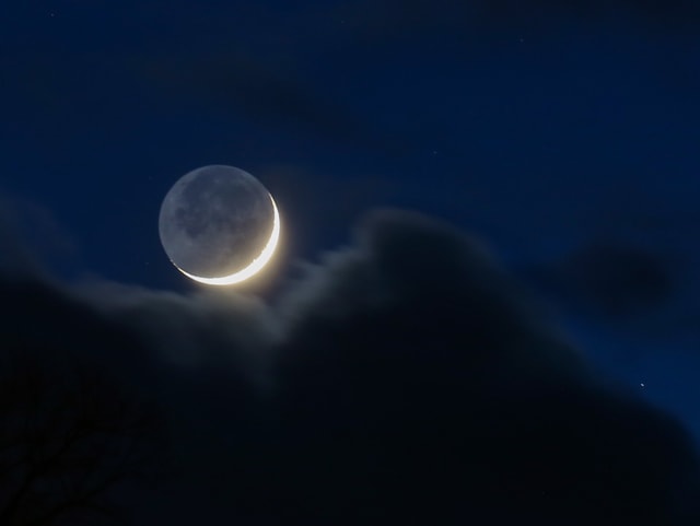 Nachthimmel mit einigen Wolken. In einer Lücke Sicht auf den Mond, von dem nur eine Sichel erhellt ist.