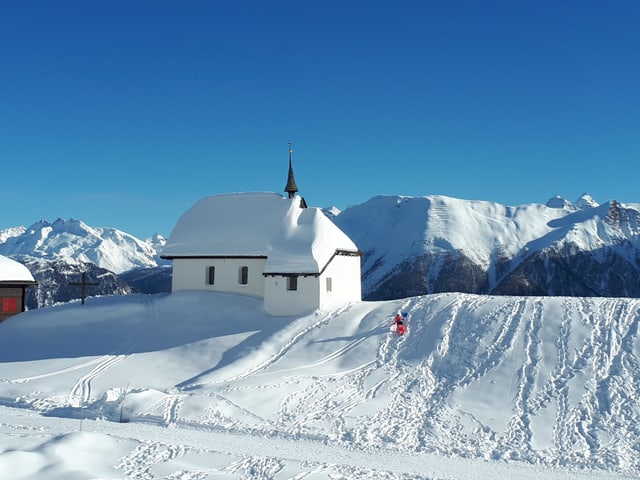 Winterlandschaft mit Kapelle auf einem Bergücke, überall liegt Schnee, der Himmel ist wolkenlos und blau.