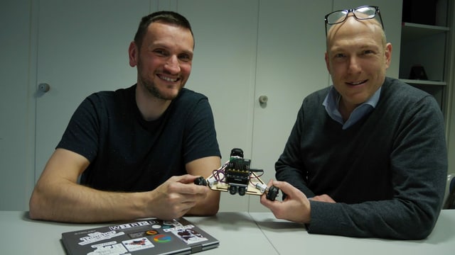 Zwei Männer sitzen an einem Tisch und halten einen kleinen Roboter in den Händen Zwei Männer sitzen an einem Tisch und halten einen kleinen Roboter in den Händen