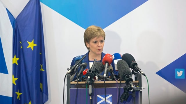 Sturgeon steht an einem Rednerpult und spricht in Mikrofone, daneben die schottische und die EU-Flagge. Sturgeon steht an einem Rednerpult und spricht in Mikrofone, daneben die schottische und die EU-Flagge.