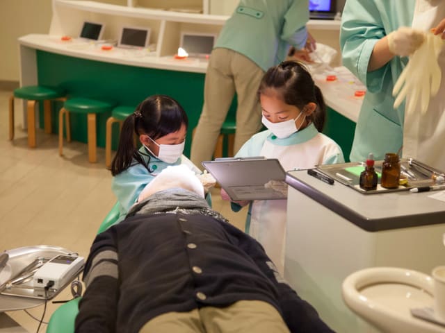 Zwei Mädchen mit Arztkitteln und Mundschutz sind in einer Zahnarztpraxis über eine liegende Puppe gebeugt. 