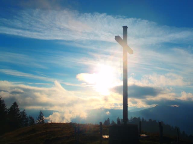 Leicht bewölkter Himmel über einem Gipfelkreuz bei Sonnenaufgang.