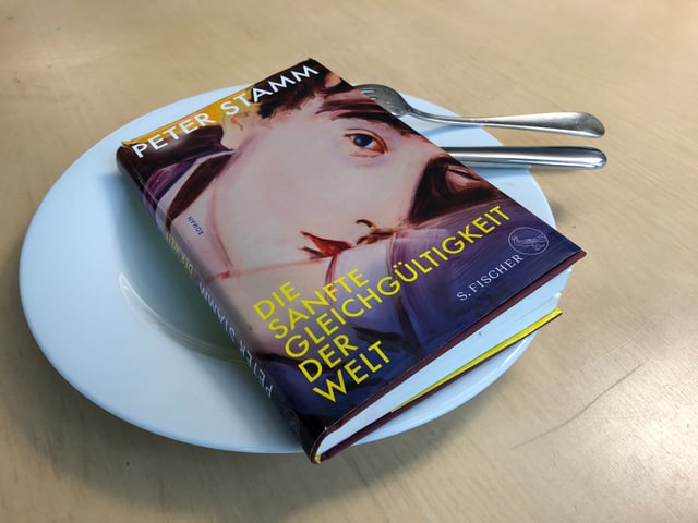 Das Buch «Die sanfte Gleichgültigkeit der Welt» von Peter Stamm liegt auf einem weissen Teller. Messer und Gabel sind hineingesteckt. Das Buch «Die sanfte Gleichgültigkeit der Welt» von Peter Stamm liegt auf einem weissen Teller. Messer und Gabel sind hineingesteckt.