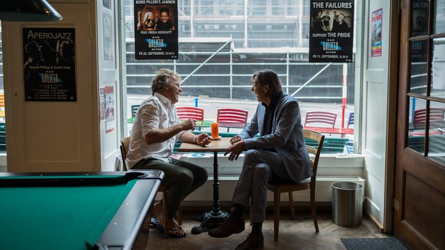 Alex Capus und Pedro Lenz diskutieren an eine Tisch in einer Bar, an der Wand hängen Plakate mit Konzerthinweisen Alex Capus und Pedro Lenz diskutieren an eine Tisch in einer Bar, an der Wand hängen Plakate mit Konzerthinweisen