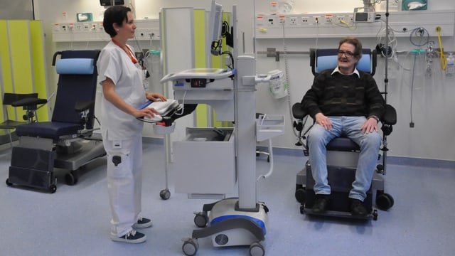 Ein Mann sitzt in einem Behandlungssessel und eine Pflegende steht an einem Computerwagen. Ein Mann sitzt in einem Behandlungssessel und eine Pflegende steht an einem Computerwagen.
