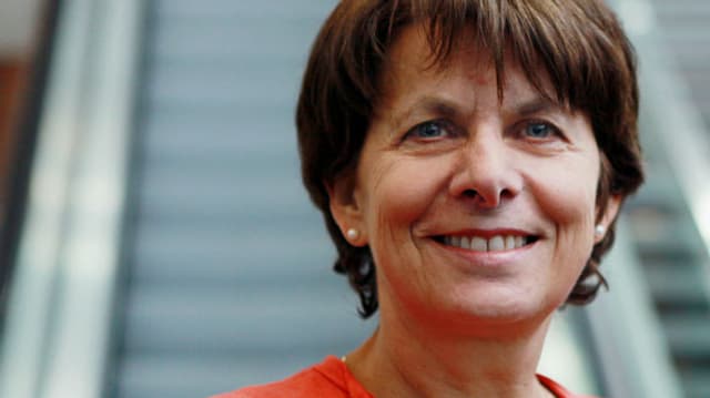 Urech hat braune, kurze Haare, blaue Augen, trägt Ohrringe und ein oranges Oberteil. Sie steht vor einer Rolltreppe. Urech hat braune, kurze Haare, blaue Augen, trägt Ohrringe und ein oranges Oberteil. Sie steht vor einer Rolltreppe.