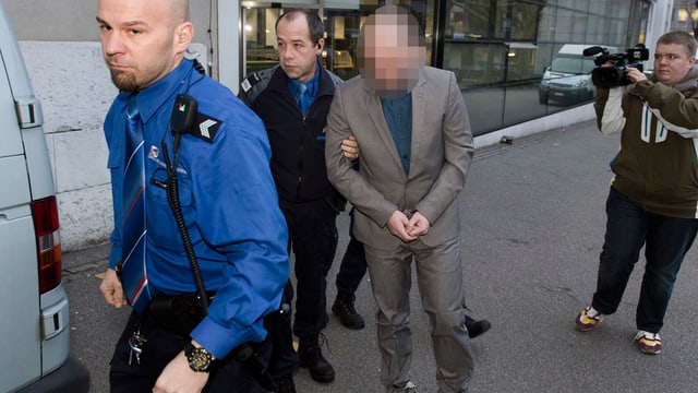 Polizisten führen einen Angeklagten zum Prozess, begleitet von einem Kameramann. Polizisten führen einen Angeklagten zum Prozess, begleitet von einem Kameramann.