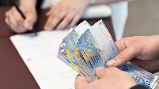 Eine Person unterschreibt ein Papier, während eine andere Person Geldnoten in den Händen hält. Eine Person unterschreibt ein Papier, während eine andere Person Geldnoten in den Händen hält.