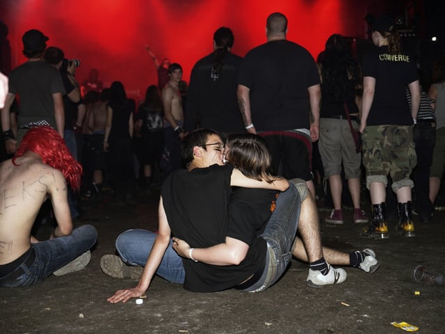 Ein Paar sitzt auf dem Boden und küsst sich, davor das Publikum von hinten während einem Konzert. 