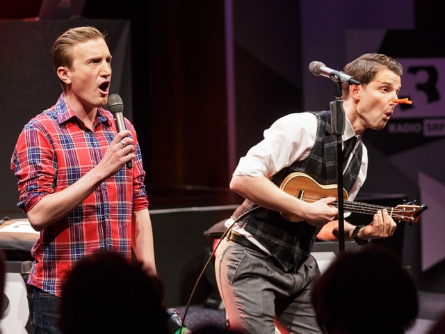 Die beiden Moderatoren auf der Bühne mit einer kleinen Gitarre Die beiden Moderatoren auf der Bühne mit einer kleinen Gitarre