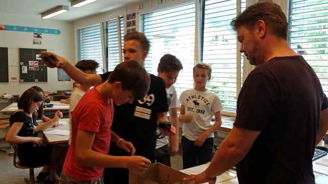 Schüler und Lehrer stehen um eine Kartonkiste, die auf einem Tisch steht und nehmen ihre Handys in Empfang. Schüler und Lehrer stehen um eine Kartonkiste, die auf einem Tisch steht und nehmen ihre Handys in Empfang.