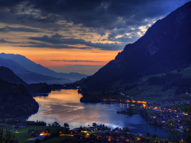 See glitzert in der Dämmerung. Rechts und links bewaldete Berghänge. Der Himmel am Horizont ist gelb bis orange und leicht bewölkt.