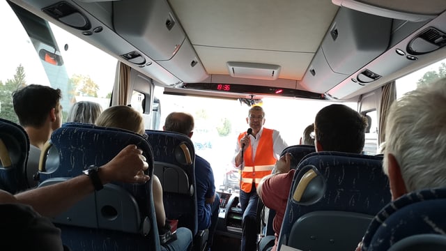 Mann in oranger Warnweste spricht in einem Bus in ein Mikrofon. Mann in oranger Warnweste spricht in einem Bus in ein Mikrofon.