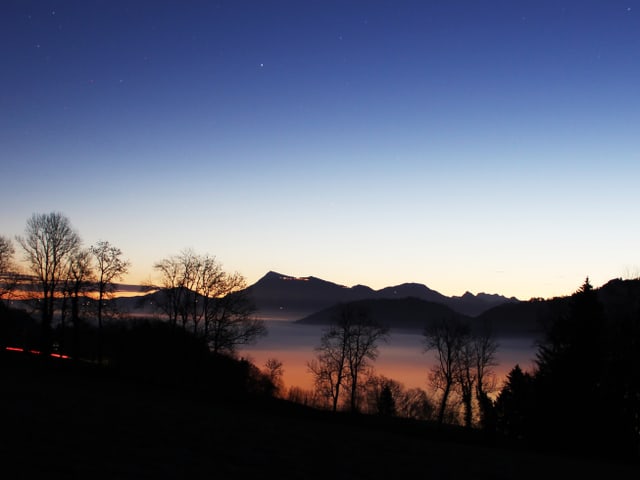 Am Horizont macht sich die Morgendämmerung bemerkbar. Der Himmel ist praktisch wolkenlos, während im Tal dichter Nebel liegt. Im Nebel sind die Lichtkegel von Dörfer und Städten zu sehen.