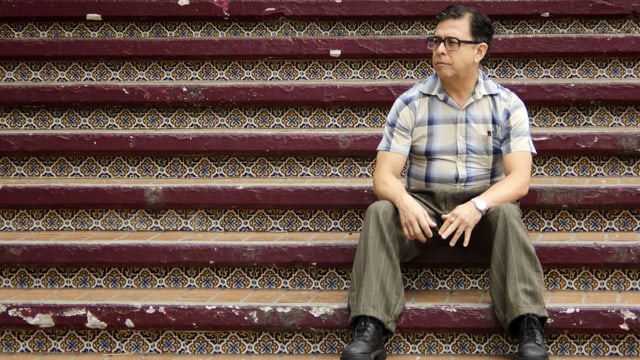 Ein älterer Mann mit dunklen Haaren, schwarzer Brille und kariertem Hemd sitzt auf einer Treppe. Ein älterer Mann mit dunklen Haaren, schwarzer Brille und kariertem Hemd sitzt auf einer Treppe.