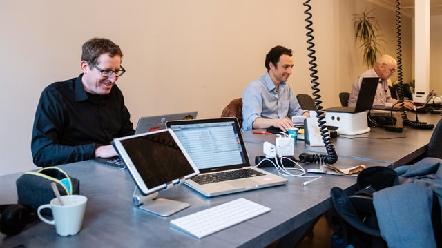Drei Männer sitzen an einem langen Tisch vor ihren Laptops. Drei Männer sitzen an einem langen Tisch vor ihren Laptops.