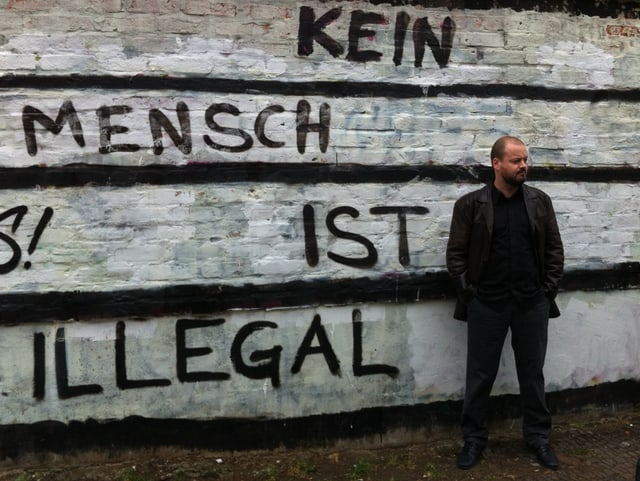 Ville Tietäväinen im Porträt. Hinten eine Mauer mit der Aufschrift «Kein Mensch ist illegal». Ville Tietäväinen im Porträt. Hinten eine Mauer mit der Aufschrift «Kein Mensch ist illegal».