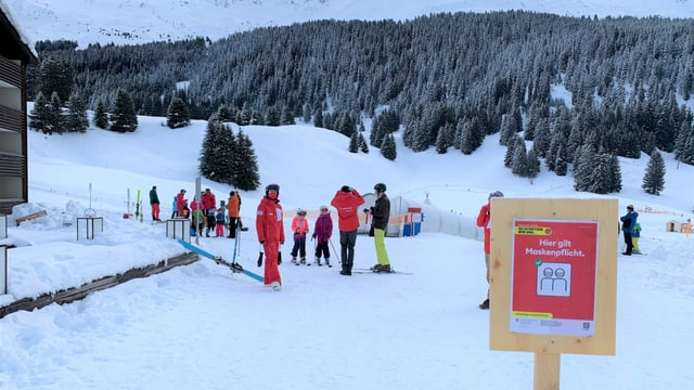 Schild "Maskenpflicht" im Schnee, im Hintergrund eine Skischulklasse