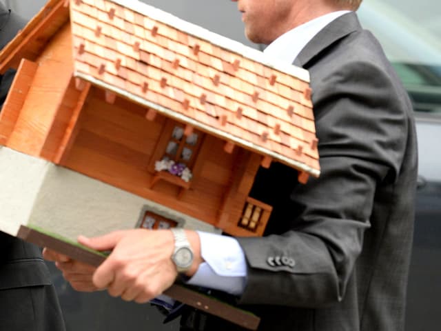 Zwei Männer, einer trägt ein Miniaturchalet. Zwei Männer, einer trägt ein Miniaturchalet.