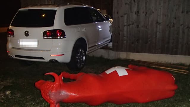 Eine rote Plastikkuh liegt am Boden - im Hintergrund steckt ein weisses Auto in einer Fassade aus Holz Eine rote Plastikkuh liegt am Boden - im Hintergrund steckt ein weisses Auto in einer Fassade aus Holz