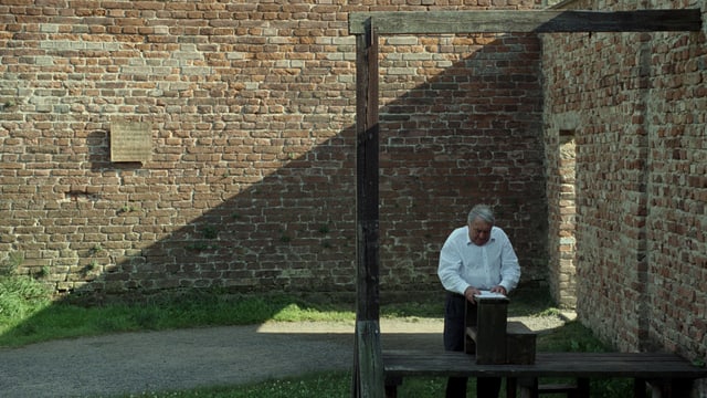 Claude Lanzmann steht unter einem Holzgebilde - dem Galgen von Theresienstadt. Im Hintergrund eine Mauer mit einer Gedenktafel. Claude Lanzmann steht unter einem Holzgebilde - dem Galgen von Theresienstadt. Im Hintergrund eine Mauer mit einer Gedenktafel.