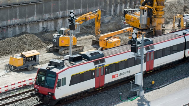 Ein Zug fährt an einer Baustelle vorbei. IM Hintergrund sind gelbe Baustellen-Fahrzeuge zu sehen. Ein Zug fährt an einer Baustelle vorbei. IM Hintergrund sind gelbe Baustellen-Fahrzeuge zu sehen.