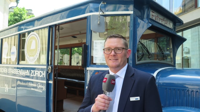 Ein Mann mit Mikrofon steht vor einem blauen, alten Bus Ein Mann mit Mikrofon steht vor einem blauen, alten Bus