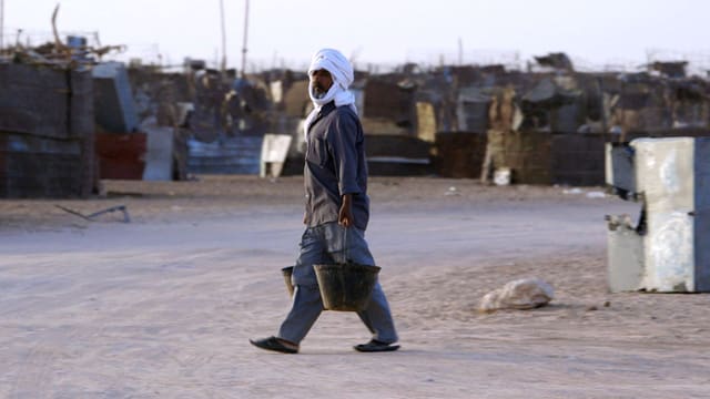Ein Mann mit Turban schleppt Wassereimer durch eine Strasse mit zerfallenen Hütten. Ein Mann mit Turban schleppt Wassereimer durch eine Strasse mit zerfallenen Hütten.