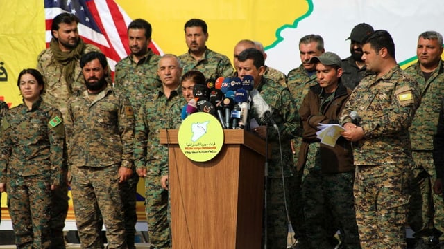Mazlum Abdi umgeben von Militär-Angehörigen am Rednerpult. Mazlum Abdi umgeben von Militär-Angehörigen am Rednerpult.