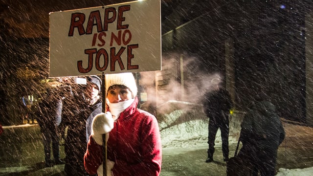 Eine Frau in einem roten Mantel hält ein weisses Transparent in der Hand. Darauf ist geschrieben: «Rape is no Joke». Eine Frau in einem roten Mantel hält ein weisses Transparent in der Hand. Darauf ist geschrieben: «Rape is no Joke».