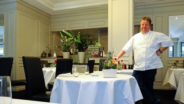Ein stämmiger Mann in weisser Kochschürze steht an einem Tisch mit weissem Tischtuch und schwarzen Stühlen und schaut in die Kamera. Ein stämmiger Mann in weisser Kochschürze steht an einem Tisch mit weissem Tischtuch und schwarzen Stühlen und schaut in die Kamera.
