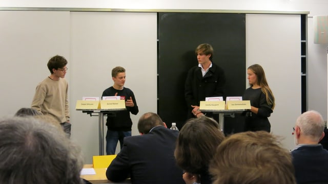 Vier Jugendliche stehen in einem Schulzimmer an Stehtischen, im Vordergrund Köpfe von Publikum und Jury. Vier Jugendliche stehen in einem Schulzimmer an Stehtischen, im Vordergrund Köpfe von Publikum und Jury.
